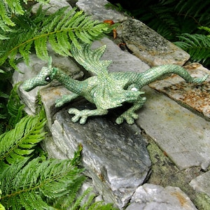May include: A green plush dragon with a textured, scaly design. The dragon is posed with its wings spread and its head turned to the side. It is sitting on a stone surface with green ferns in the background.