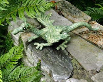 May include: A green plush dragon with a textured, scaly design. The dragon is posed with its wings spread and its head turned to the side. It is sitting on a stone surface with green ferns in the background.
