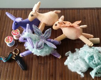 Könnte beinhalten: Drei handgefertigte Plüschtiere in Tierform. Das erste ist ein lila-blaues Einhorn mit einer hellblauen, flauschigen Mähne. Das zweite ist ein helloranger Hirsch mit einem hellbraunen Körper. Das dritte ist ein hellrosa Hirsch mit einem hellbraunen Körper. Die Spielzeuge liegen auf einer braunen Bambusmatte.