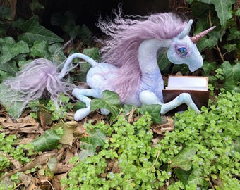 Könnte beinhalten: Ein skurriles Plüsch-Einhorn mit lavendelfarbener Mähne und Schweif, blauem Körper und goldenem Horn. Das Einhorn liegt inmitten von grünem Laub und scheint ein kleines, offenes Buch zu lesen. Die Gesamtästhetik ist Fantasy-Themen.