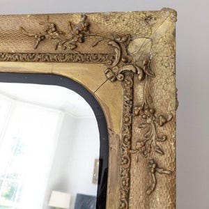 May include: A rectangular wall mirror featuring a gold-toned frame with a textured, stippled surface. The frame is adorned with raised floral and scrollwork carvings at the corners and edges, showing visible signs of age and minor cracks.