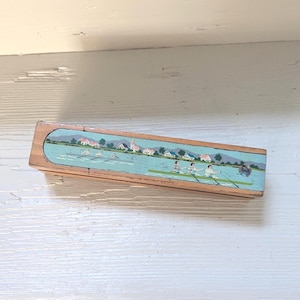 Peut inclure: Une boîte rectangulaire en bois avec un couvercle coulissant orné d'une scène peinte représentant une équipe d'aviron sur une rivière. L'illustration montre des rameurs dans un long bateau, une rive avec des maisons, des arbres et un clocher.
