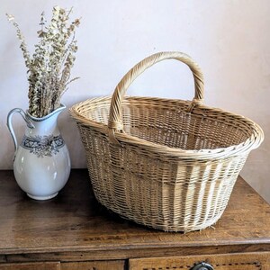 May include: A large, oval wicker basket with a curved handle, displayed on a wooden surface. The basket is a natural light brown colour. A white jug with a floral pattern and dried flowers is positioned to the left of the basket.