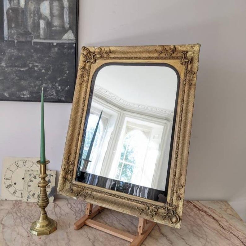 May include: A rectangular wall mirror featuring an ornate, textured gold frame with decorative corner detailing. The mirror is supported by a wooden easel stand. The reflective surface shows a clear view of a room with windows and natural light.