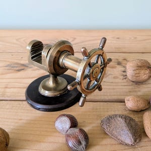 May include: A vintage brass nutcracker shaped like a ship's wheel, mounted on a black wooden base. The nutcracker has a detailed design and is surrounded by various nuts on a wooden surface.
