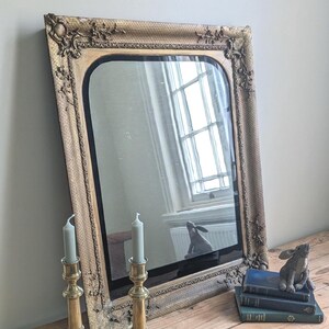 May include: A rectangular wall mirror with a gold-toned, ornate frame featuring raised floral and scrollwork details. The mirror has a smooth, reflective surface with rounded top corners, reflecting a window and a small rabbit figurine.