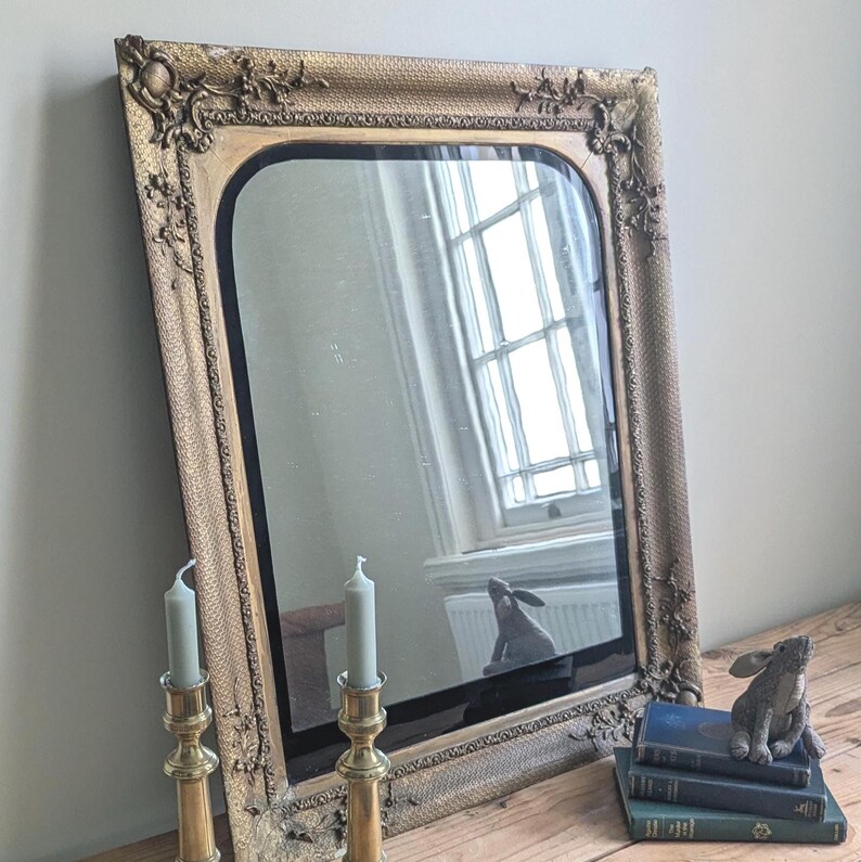 May include: A rectangular wall mirror with a gold-toned, ornate frame featuring raised floral and scrollwork details. The mirror has a smooth, reflective surface with rounded top corners, reflecting a window and a small rabbit figurine.