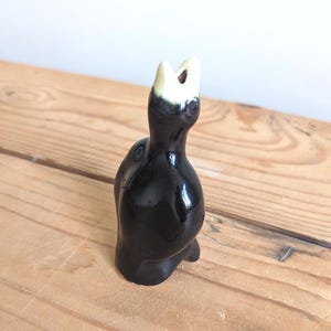 May include: A small, glossy black ceramic figurine of a bird with its head tilted upwards and its beak open. The bird features a cream-coloured beak and head top, with a smooth, reflective surface finish and a rounded body shape standing upright.