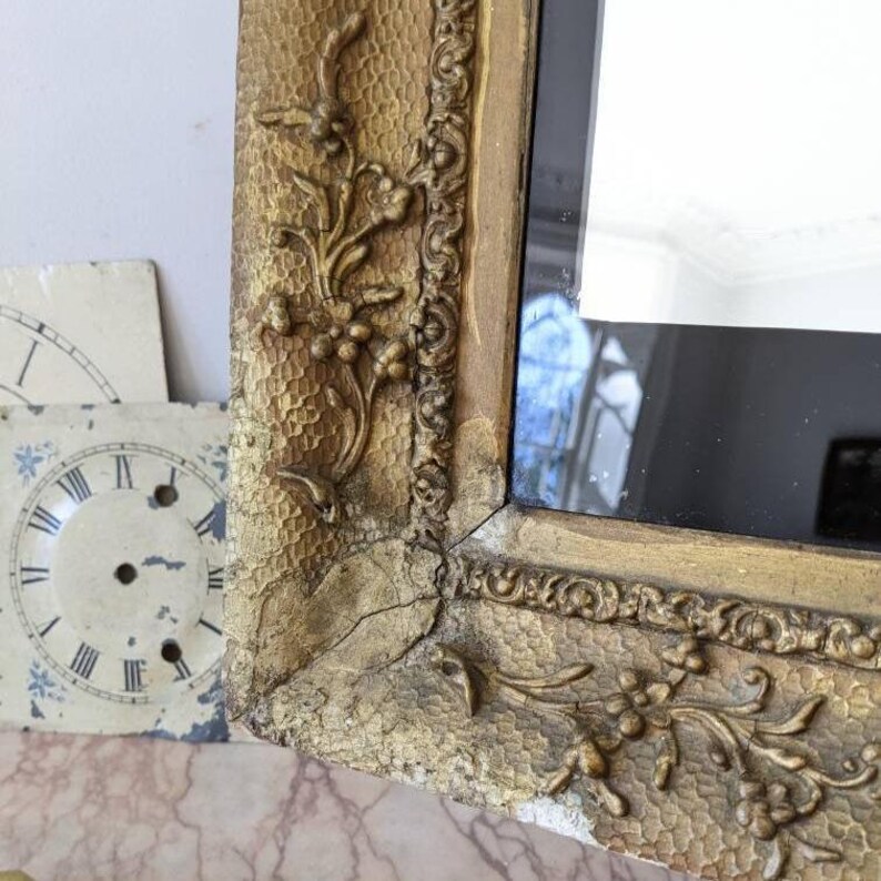 May include: A gold-toned rectangular mirror frame featuring intricate relief carvings of floral vines and a textured, dimpled surface. The frame shows visible signs of age, including cracks and worn areas, surrounding a reflective glass surface.
