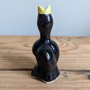 May include: A small, glossy black ceramic figurine of a bird with a bright yellow beak. The bird is depicted in a standing position with its head tilted upward, showcasing a smooth, reflective surface and a simple, stylised form.
