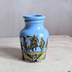 Puede incluir: Un tarro de cerámica azul claro con un diseño decorativo que presenta figuras en un círculo alrededor de un reloj de arena con el texto "All the world's a stage".