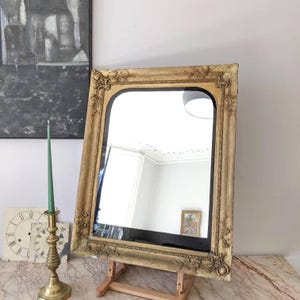 May include: A rectangular mirror with a gold-toned, ornate frame featuring raised floral and scrollwork details. The mirror has rounded upper corners and is supported by a wooden easel stand. A tall green taper candle in a brass holder sits nearby.