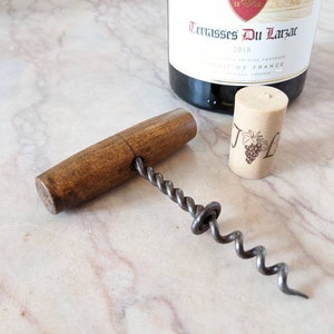Puede incluir: Un sacacorchos de mango de madera con una espiral de metal. El sacacorchos está sobre una superficie de mármol blanco. También se puede ver un corcho de una botella de vino en la imagen.