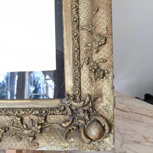 May include: A rectangular mirror featuring an ornate gold-toned frame with a textured, stippled surface. The frame is decorated with raised floral carvings and a beaded inner border, with a prominent rounded corner detail and visible signs of wear.