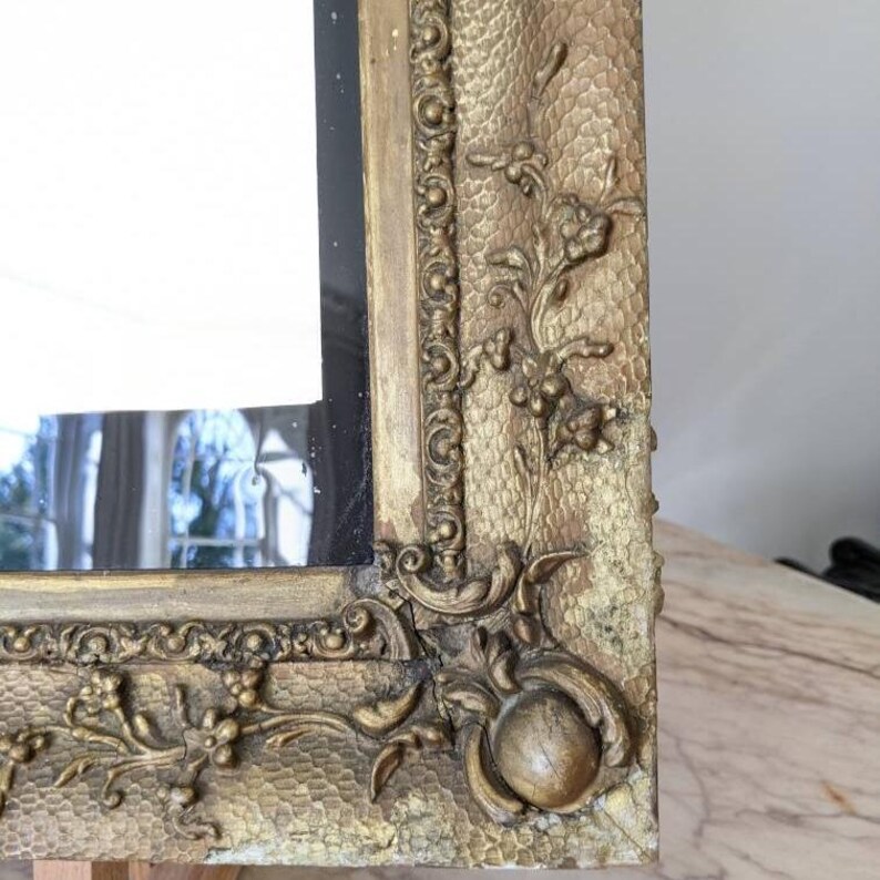 May include: A rectangular mirror featuring an ornate gold-toned frame with a textured, stippled surface. The frame is decorated with raised floral carvings and a beaded inner border, with a prominent rounded corner detail and visible signs of wear.