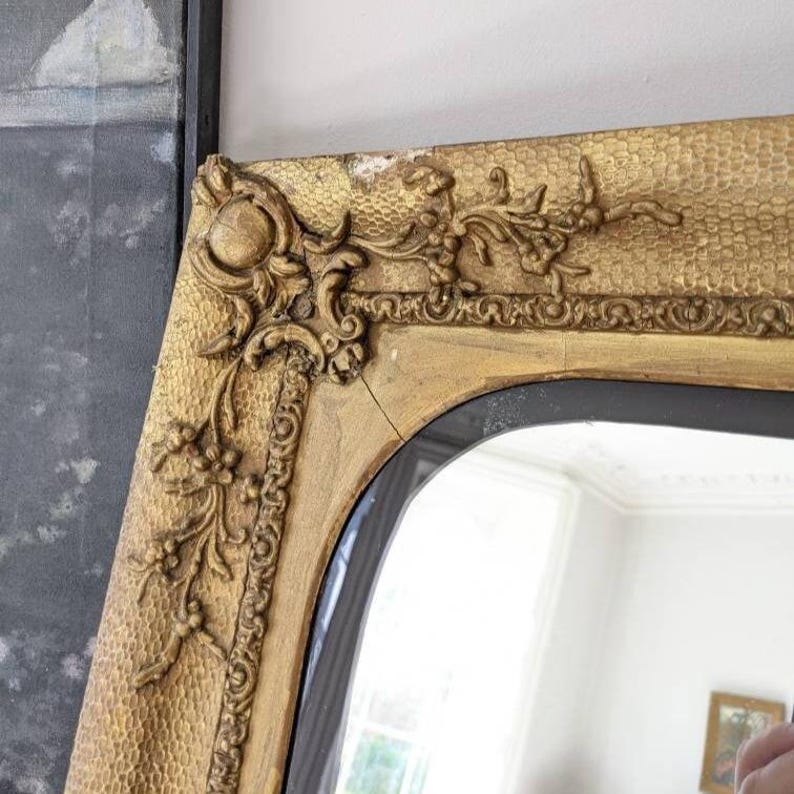 May include: A gold-toned mirror featuring an ornate, textured frame with raised floral and scrollwork detailing at the corner. The frame exhibits a hammered surface finish and a visible crack along the upper section of the wooden moulding.