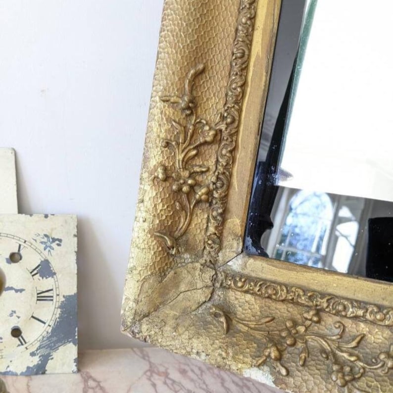 May include: A rectangular mirror with a gold-toned frame featuring a textured, hammered surface and raised floral relief carvings. The frame displays visible signs of wear, including cracks and areas where the gold finish has faded.