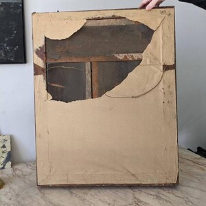 May include: The back of a rectangular picture frame featuring a dark brown wooden border. A large, torn piece of light tan paper covers the back, revealing a dark brown wooden panel underneath. A thin metal wire is attached for hanging purposes.