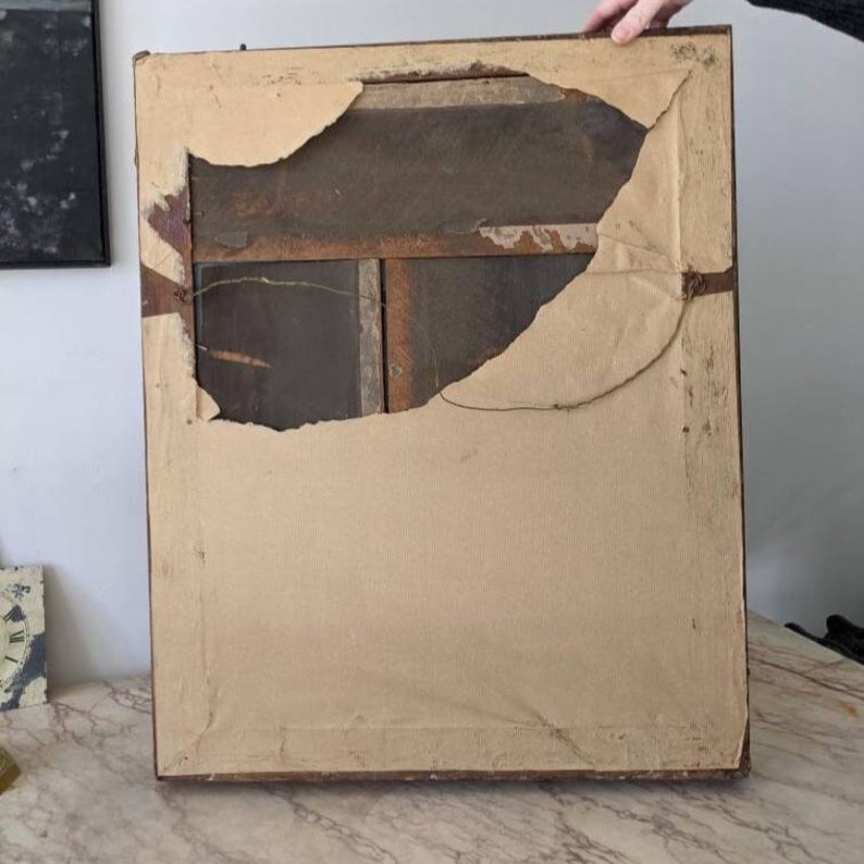 May include: The back of a rectangular picture frame featuring a dark brown wooden border. A large, torn piece of light tan paper covers the back, revealing a dark brown wooden panel underneath. A thin metal wire is attached for hanging purposes.