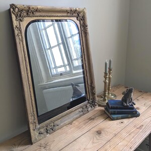 May include: A rectangular wall mirror with a gold-toned frame featuring ornate floral and scrollwork relief details at the corners. The mirror has a rounded top edge and reflects a window and a small rabbit figurine sitting on a stack of books.