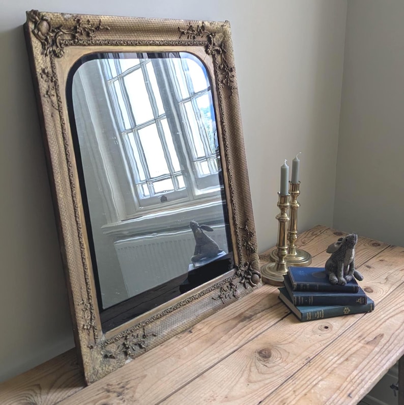 May include: A rectangular wall mirror with a gold-toned frame featuring ornate floral and scrollwork relief details at the corners. The mirror has a rounded top edge and reflects a window and a small rabbit figurine sitting on a stack of books.