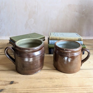 May include: Two brown stoneware mugs with a glazed finish. The mugs have a rounded body and a handle. The mugs are sitting on a wooden surface.