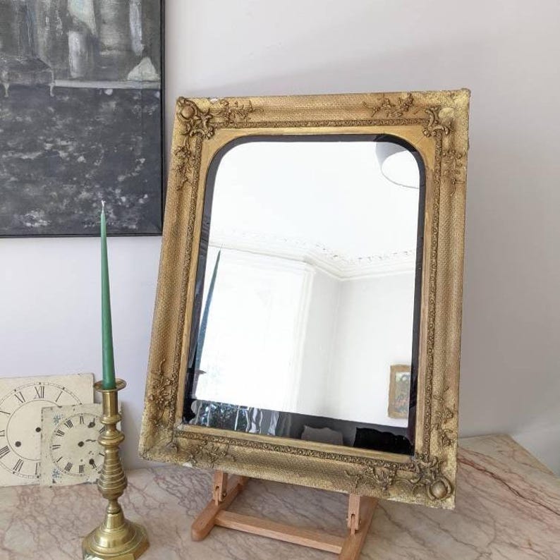 May include: A rectangular mirror with a gold-toned frame featuring ornate, raised floral and scrollwork detailing at the corners and edges. The mirror has a smooth, reflective surface and is supported by a wooden stand, displaying a clear reflection.
