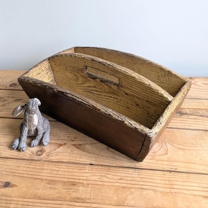 May include: A rectangular wooden caddy with a central divider and a handle cutout, featuring a dark brown exterior and a lighter, weathered yellow interior finish. A small, grey, patterned fabric rabbit figurine sits beside the wooden caddy.