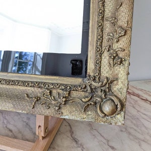 May include: A rectangular mirror featuring a wide, gold-toned frame with a textured, hammered surface. The frame is decorated with raised floral and scrollwork relief patterns at the corners and edges, showcasing a metallic finish with aged details.