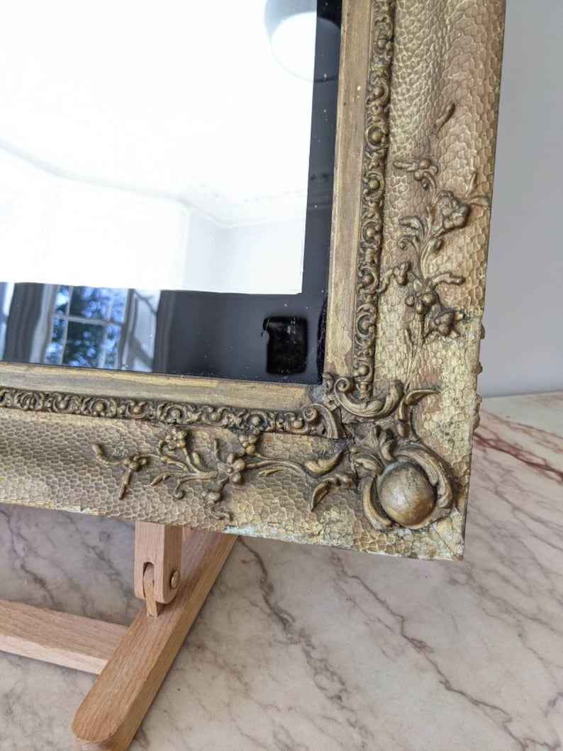 May include: A rectangular mirror featuring a wide, gold-toned frame with a textured, hammered surface. The frame is decorated with raised floral and scrollwork relief patterns at the corners and edges, showcasing a metallic finish with aged details.
