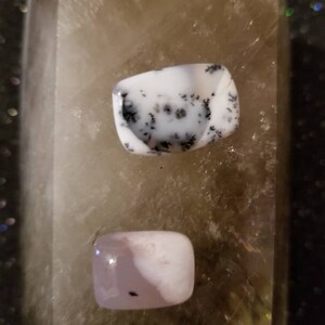 May include: Two polished rectangular gemstones resting on a larger translucent crystal. The top stone is opaque white with black dendritic fern-like inclusions. The bottom stone is a smooth, pale pinkish-white cabochon with a small dark speck.