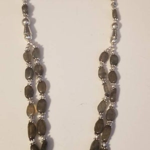 Peut inclure: Un collier à plusieurs rangs composé de pierres grises lisses de forme ovale, alternées avec de petites perles argentées rondes. Le design comprend une perle centrale cylindrique en métal argenté ornée reliant les deux rangs de pierres.