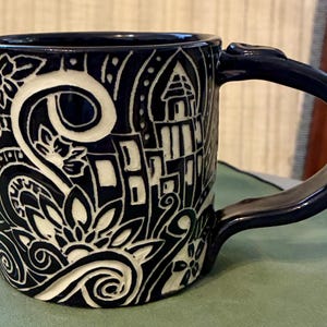 Könnte beinhalten: Eine marineblaue Keramiktasse mit einem weißen, geschnitzten Design mit floralen und architektonischen Motiven. Die Tasse hat einen gebogenen Henkel und eine dunkelblaue Innenseite. Das Design ist ein Reliefmuster, das eine strukturierte Oberfläche erzeugt.