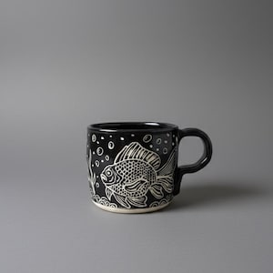 May include: Black ceramic mug featuring a white goldfish illustration, bubbles, and aquatic plants. The mug has a rounded handle and a white base. The design is hand-drawn.