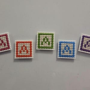 May include: Five square, cross-stitched magnets. Each magnet features a white letter "A" on a colored background. The borders are pink, orange, green, blue, and purple. The magnets are arranged on a light gray surface.