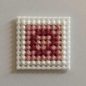 Puede incluir: Una pieza cuadrada de punto de cruz con un borde blanco y un diseño central en tonos rojos y rosas. La costura crea un patrón texturizado, mostrando el arte de la aguja. La pieza es probablemente un pequeño objeto decorativo.