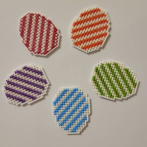 May include: Five colorful, striped, oval-shaped coasters. The coasters are made of white fabric with a cross-stitch pattern. The colors are red, orange, purple, green, and blue.