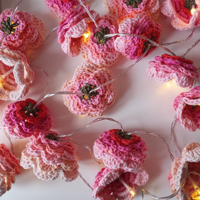 Op de afbeelding: Een slinger van roze en rode gehaakte bloemenlampjes. De lampjes zijn aan elkaar geregen met een doorzichtig draad en elke bloem heeft een klein LED-lampje.