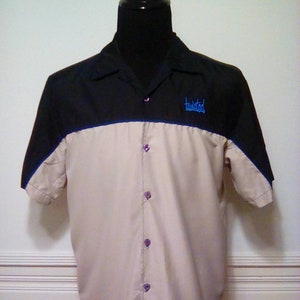May include: A short-sleeved button-up shirt with a black upper section and tan lower section. The shirt has a blue trim and the word "twisted" embroidered in blue. The buttons are purple.