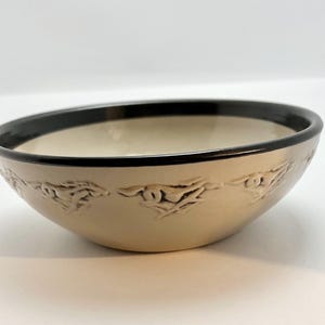May include: A beige ceramic bowl with a black rim. The bowl features a series of embossed, stylized horse designs around the exterior. The interior of the bowl is a lighter beige color.
