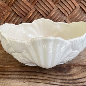 May include: A cream-colored, shell-shaped ceramic bowl. The bowl has a scalloped rim and a textured, speckled surface. The bowl is sitting on a wooden surface. The background is a woven wooden pattern.