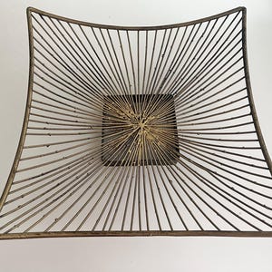 May include: A square-shaped decorative bowl constructed from thin, dark bronze-toned metal wires that radiate outward from a solid, square-shaped center base, creating a sunburst pattern with a textured, weathered finish.