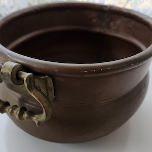 May include: A copper planter with a decorative brass handle. The planter is round and has a slightly flared lip.