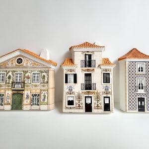 May include: Three ceramic house-shaped containers with orange tiled roofs. The left house features a green door and ornate painted details. The center house has three levels with black balconies. The right house displays a blue and white tile pattern.