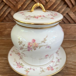 May include: A white porcelain jar with a lid and matching saucer. The jar and lid feature a floral pattern in pink and yellow, with gold trim. The lid has a gold handle. The set is on a wooden surface.
