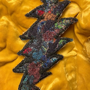 May include: A decorative, multi-colored lightning bolt applique with a black border, set against a golden yellow velvet background. The applique features a patchwork design with various colors and textures.
