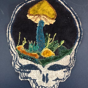Könnte beinhalten: Gestickter Aufnäher mit einem Totenkopf-Design, das eine Pilz- und Pflanzenszene zeigt. Der Totenkopf ist grau mit blauen Akzenten, und der Pilz hat einen gelben Hut und einen blauen Stiel. Der Hintergrund ist dunkelblauer Cord.