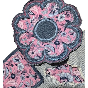 May include: A denim fabric patch shaped like a flower with scalloped edges, featuring a pink and blue floral pattern. The patch is stitched onto a light blue denim base, which also includes a square patch and a distressed area with frayed edges.