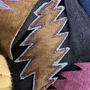 May include: Close-up of colorful corduroy fabric pieces with embroidered lightning bolt designs. The bolts are brown with blue and purple accents, set against a black background. Other fabric pieces are in shades of teal, brown, and purple.