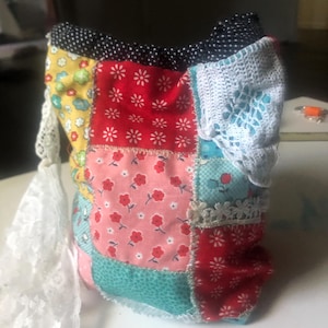 May include: A patchwork bag with a variety of colorful floral and polka dot patterns. The bag features a white lace trim and a white lace strap. The bag is made of fabric and has a handmade appearance.